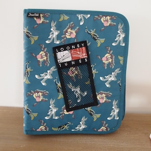 May include: Teal binder with a Looney Tunes design. The binder features a repeating pattern of Bugs Bunny and Taz. The binder has a mesh pocket and the words "Looney Tunes" are visible on a tag.