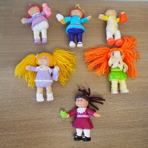 May include: A collection of six vintage Cabbage Patch Kids miniature dolls. The dolls have various hair colors, including yellow and orange, and wear colorful outfits. Some dolls are holding accessories.