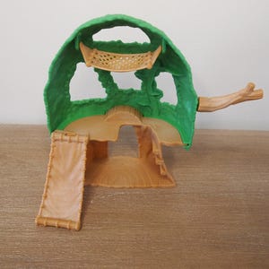May include: A green and tan plastic playset resembling a treehouse. The playset features a green canopy, a tan base with a ramp, and a small branch extending from the side. The playset is designed for children's imaginative play.