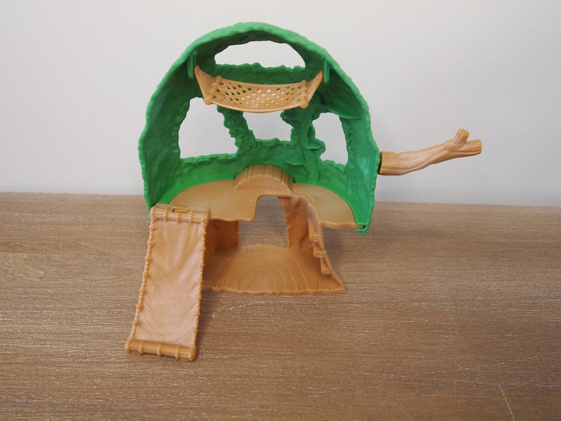 May include: A green and tan plastic playset resembling a treehouse. The playset features a green canopy, a tan base with a ramp, and a small branch extending from the side. The playset is designed for children's imaginative play.