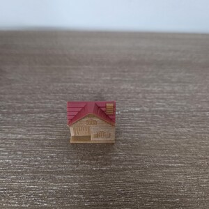 Pode incluir: Uma figura de casa em miniatura com telhado vermelho, paredes bege e uma pequena chamin&eacute;. A casa tem uma porta da frente e janelas. A figura est&aacute; sobre um fundo de madeira.