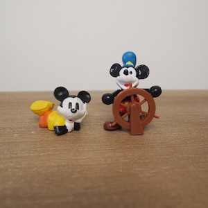 May include: Two Mickey Mouse figurines. One is crawling, wearing yellow and orange clothing. The other is standing, dressed as a captain with a blue hat, holding a brown ship's wheel. Both figures have black ears and white faces.
