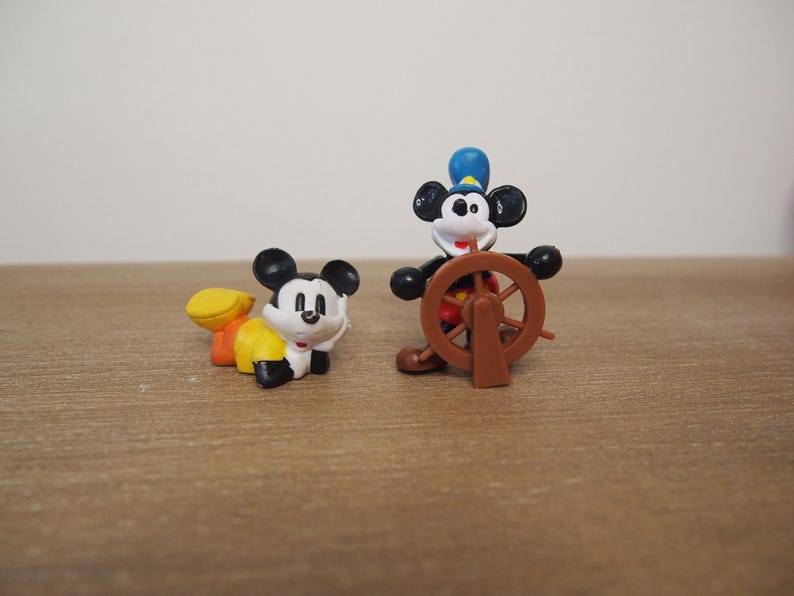 May include: Two Mickey Mouse figurines. One is crawling, wearing yellow and orange clothing. The other is standing, dressed as a captain with a blue hat, holding a brown ship's wheel. Both figures have black ears and white faces.
