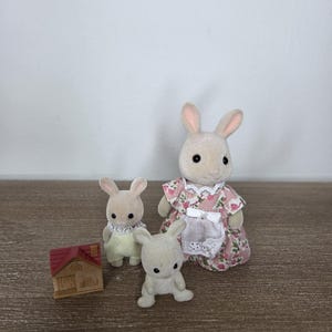 Pode incluir: Uma fam&iacute;lia de coelhos de brinquedo, incluindo uma m&atilde;e com um vestido floral, uma crian&ccedil;a com um vestido amarelo e um coelho sentado. Uma casa de madeira em miniatura com um telhado vermelho tamb&eacute;m est&aacute; presente. Os brinquedos s&atilde;o bege e brancos.
