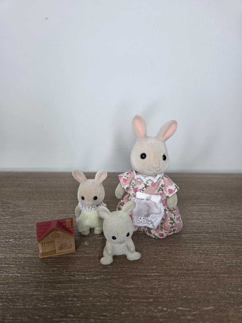Pode incluir: Uma fam&iacute;lia de coelhos de brinquedo, incluindo uma m&atilde;e com um vestido floral, uma crian&ccedil;a com um vestido amarelo e um coelho sentado. Uma casa de madeira em miniatura com um telhado vermelho tamb&eacute;m est&aacute; presente. Os brinquedos s&atilde;o bege e brancos.