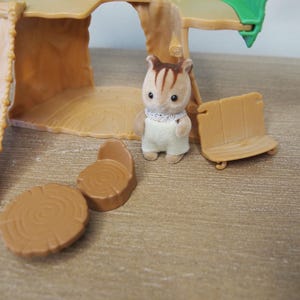 May include: A miniature toy scene featuring a small brown and white squirrel figure wearing a white bib and overalls. The scene includes a wooden bench, two tree stump tables, and a treehouse structure. The toys are made of plastic and are set on a wooden surface.