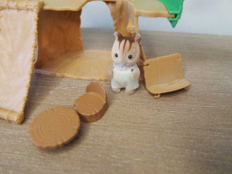 May include: A miniature toy scene featuring a small brown and white squirrel figure wearing a white bib and overalls. The scene includes a wooden bench, two tree stump tables, and a treehouse structure. The toys are made of plastic and are set on a wooden surface.