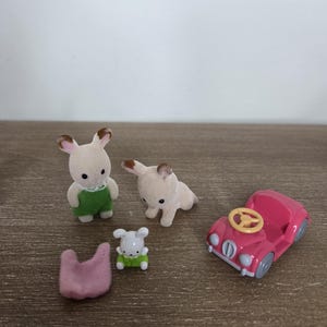 May include: A collection of miniature toy figures and accessories. Includes two bunny figures, a dog figure, a small car, and a pink felt item. The bunny figures wear green overalls. The car is pink with a yellow steering wheel.