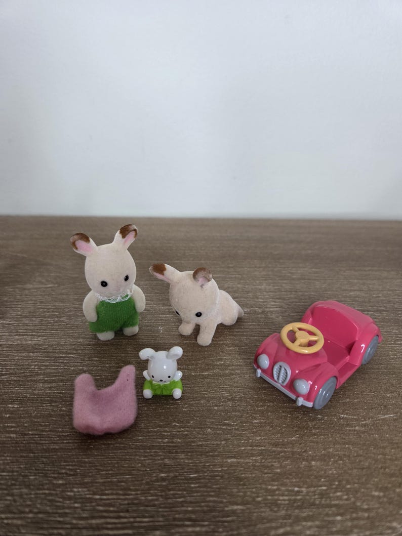 May include: A collection of miniature toy figures and accessories. Includes two bunny figures, a dog figure, a small car, and a pink felt item. The bunny figures wear green overalls. The car is pink with a yellow steering wheel.