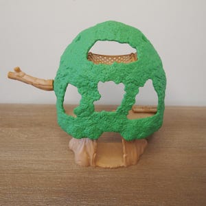 May include: A green plastic treehouse toy with a textured surface. The treehouse has openings and a small branch extending to the left. The base and branch are tan. The toy is on a wooden surface.