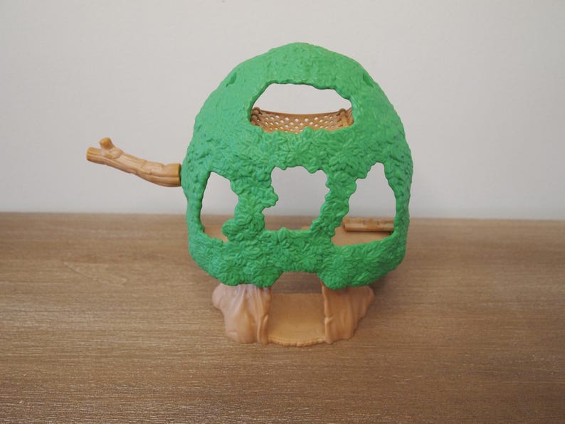 May include: A green plastic treehouse toy with a textured surface. The treehouse has openings and a small branch extending to the left. The base and branch are tan. The toy is on a wooden surface.