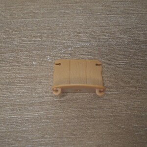 May include: A small, tan-colored decorative shelf with a scroll design on the ends. The shelf has a flat surface and two small holes at the top for hanging. The shelf is made of a smooth material.