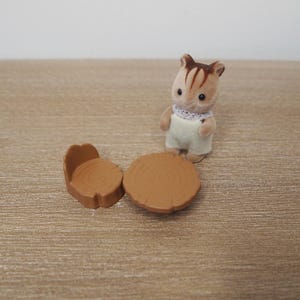 May include: A miniature toy scene featuring a brown and white chipmunk figure wearing overalls and a lace collar, standing near a small brown table and chair set. The table and chair are designed to look like tree stumps.