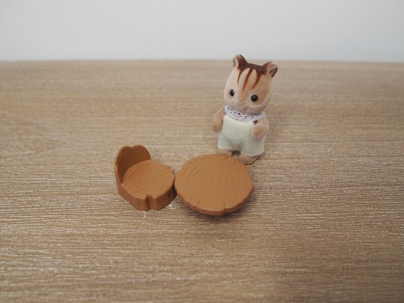 May include: A miniature toy scene featuring a brown and white chipmunk figure wearing overalls and a lace collar, standing near a small brown table and chair set. The table and chair are designed to look like tree stumps.