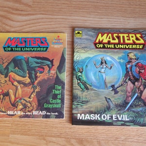 May include: Two vintage Masters of the Universe comic books. The first book is titled "The Thief of Castle Grayskull" and features a green and red character leaping over a fallen figure. The second book is titled "Mask of Evil" and features a group of characters in a swampy setting.
