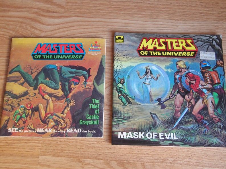 May include: Two vintage Masters of the Universe comic books. The first book is titled "The Thief of Castle Grayskull" and features a green and red character leaping over a fallen figure. The second book is titled "Mask of Evil" and features a group of characters in a swampy setting.