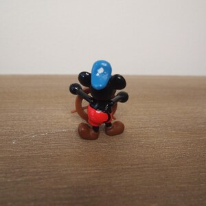 May include: A small figurine of a cartoon mouse wearing a blue hat and red shorts, holding a brown ship's wheel. The mouse has black ears and a black body. The figurine is on a brown wooden surface.