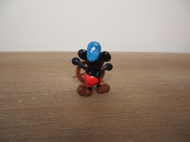 May include: A small figurine of a cartoon mouse wearing a blue hat and red shorts, holding a brown ship's wheel. The mouse has black ears and a black body. The figurine is on a brown wooden surface.