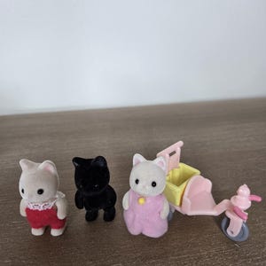 May include: Three miniature cat figurines, one black and two white, with red and pink outfits. A pink and yellow toy scooter is also present. The toys are set on a wooden surface.
