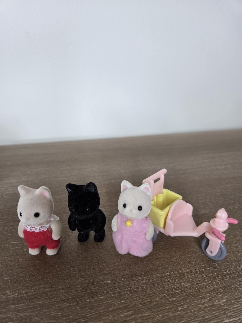 May include: Three miniature cat figurines, one black and two white, with red and pink outfits. A pink and yellow toy scooter is also present. The toys are set on a wooden surface.