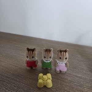 May include: Three miniature squirrel figurines wearing red, green, and pink outfits with white lace collars. A small yellow sandcastle toy is in front of the figurines. The toys are on a wooden surface.