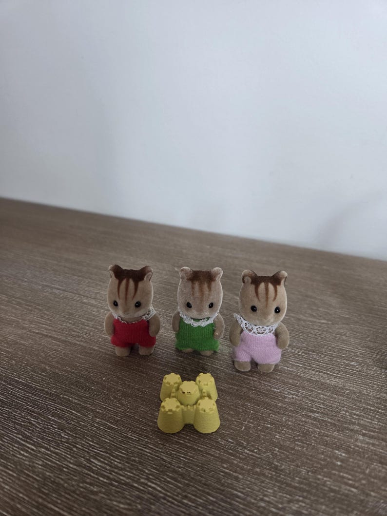 May include: Three miniature squirrel figurines wearing red, green, and pink outfits with white lace collars. A small yellow sandcastle toy is in front of the figurines. The toys are on a wooden surface.
