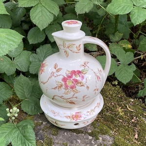 Könnte beinhalten: Ein weißes Keramik-Krug- und Waschbecken-Set mit rosa und braunen Blumenmustern. Der Krug hat einen Griff und einen Deckel. Das Waschbecken ist kreisförmig mit einem kleinen Loch. Das Set steht auf einem moosbewachsenen Felsen.