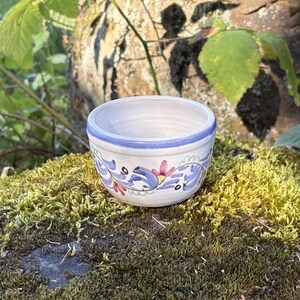 May include: A small, round ceramic bowl with a white base, featuring a hand-painted blue rim and a decorative pattern of blue swirling vines and small pink flowers encircling the exterior. The interior of the bowl is a smooth, off-white finish.