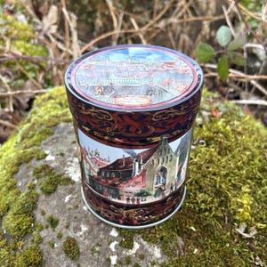 May include: A cylindrical tin with a detailed illustration of a European townscape. The tin has a dark red and black decorative pattern around the top and bottom. The lid shows a panoramic view of the town.