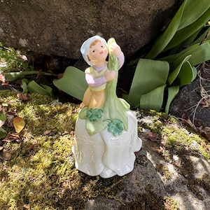 May include: A porcelain figurine of a child holding a flower. The figurine is primarily white, with accents of green, yellow, and lavender. The child wears a light blue cap and a matching lavender and yellow outfit. The figurine is set on a mossy rock.