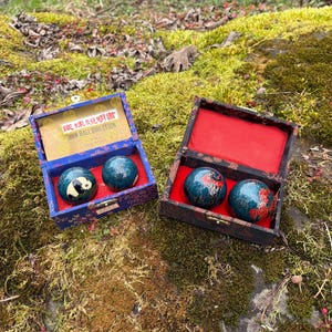 Può includere: Due set di palle di ferro cinesi in scatole decorative. Una scatola è blu con un disegno di panda su una palla. L'altra scatola è scura con fodera rossa. Le palle sono verde acqua con accenti rossi e dorati. Le scatole sono aperte.