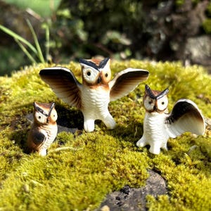 May include: Three ceramic owl figurines with brown and white coloring are displayed on a bed of green moss. One owl has its wings spread, while the other two have their wings partially open or closed. The owls are a decorative item.