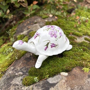 Könnte beinhalten: Eine weiße Keramik-Schildkrötenfigur mit floralem Design in Lilatönen. Die Schildkröte befindet sich auf einem moosbedeckten Felsen, der ihre dekorativen Details zeigt. Ein bezaubernder Dekorationsartikel.