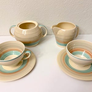 May include: A ceramic tea set featuring a cream base with horizontal bands in shades of teal, peach, and brown. The set includes a lidded sugar bowl with two handles, a creamer pitcher, and two teacups with matching saucers, all with a smooth finish.