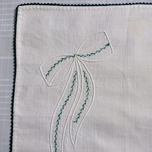 May include: White linen napkin with a green embroidered bow design. The bow is detailed with green stitching, and the edges of the napkin are trimmed with a green scalloped border. The napkin is a square shape.