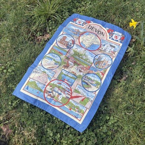 May include: A rectangular blue and white tea towel featuring a map and illustrations of Devon, England. The towel has a blue border and includes images of coastal scenes, buildings, and the word "Devon" at the top.