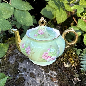 May include: A light green porcelain teapot featuring a scalloped edge, a gold-toned handle, a gold-toned spout, and a gold-toned finial on the lid. The teapot is decorated with pink rose floral patterns on the body and the lid.
