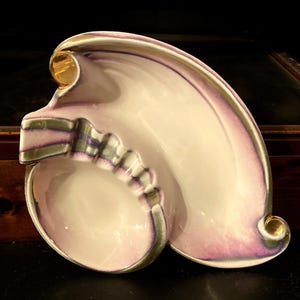 Könnte beinhalten: Ein Vintage-Aschenbecher aus Keramik mit einem einzigartigen, geschwungenen Design. Der Aschenbecher hat eine weiße Basis mit rosa und lila Akzenten und goldenen Verzierungen. Er hat eine abgerundete Schale und eine wellenförmige Form. Ein dekoratives Objekt.