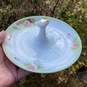 May include: A white porcelain serving dish with a green and gold rim and floral design. The dish has a handle in the center.