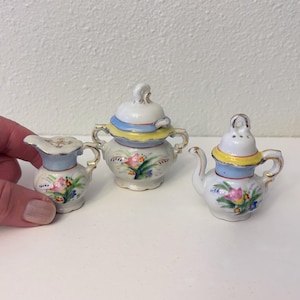 May include: A set of three miniature porcelain teacups with floral designs. The cups are white with yellow, blue, and pink accents. The cups are decorated with flowers and leaves.