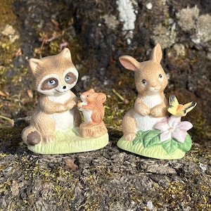 May include: Two ceramic figurines of woodland creatures. One is a raccoon with blue eyes and a squirrel. The other is a bunny with a butterfly and a flower. The figurines are painted in soft, natural colours.