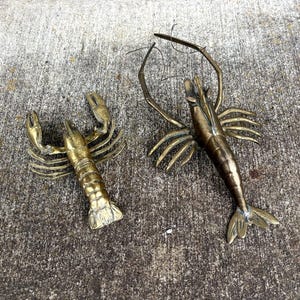 May include: Two brass crustacean figurines, a lobster and a shrimp, rest on a textured concrete surface. The lobster has large claws and a segmented body. The shrimp has long antennae and a slender body.