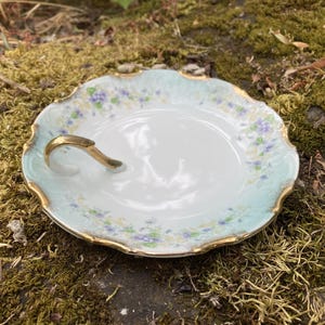 以下が含まれることがあります： 金色の取っ手と縁取りが付いた、白と淡い青色の磁器製サービング皿。紫と黄色のアクセントの花柄が特徴です。スカラップの縁が装飾的なタッチを加えています。