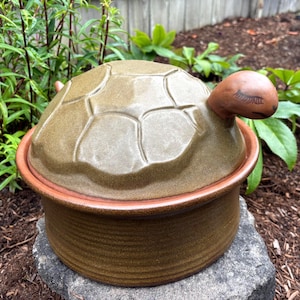 May include: A ceramic lidded container shaped like a turtle. The lid is olive green with a raised shell pattern and a brown head featuring painted closed eyes. The base is a cylindrical brown bowl with a textured surface and a smooth rim.