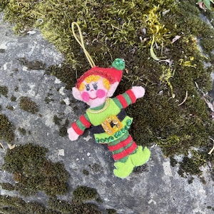 May include: A felt elf ornament featuring a red hat with a green pom-pom, yellow hair, and pink cheeks. The elf wears a green tunic with a black belt, gold sequins, and green striped sleeves and leggings, finished with green pointed shoes.