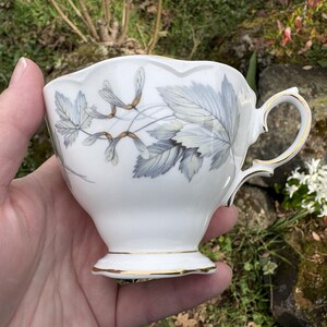 May include: White teacup with a gold rim and handle, featuring a delicate design of gray and green leaves and seed pods. The cup has a scalloped edge and sits on a small foot. A classic piece for tea or coffee.