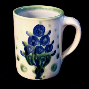 Puede incluir: Una taza de cerámica con base blanca, que presenta un motivo floral pintado a mano en azul y verde en la parte frontal. El borde está acentuado con una banda de color verde claro y la taza incluye un asa curva en el lado derecho.