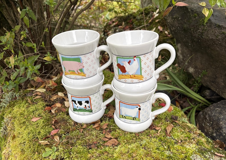 May include: Four white ceramic mugs stacked on top of each other. Each mug features a different farm animal illustration: a pig, a cow, a chicken, and a sheep. The mugs have a handle and a decorative pattern.