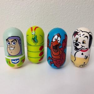 May include: Four colorful, cylindrical toys featuring cartoon characters. The toys include Buzz Lightyear, a green alien, Sebastian the crab, and a Dalmatian dog. Each toy has a unique design and color scheme, standing upright on a white surface.