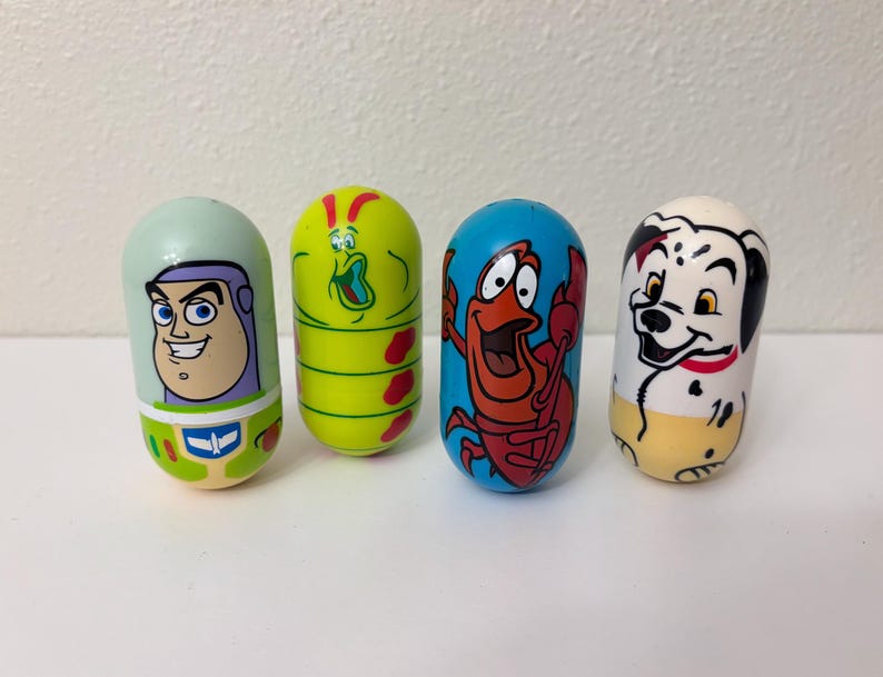 May include: Four colorful, cylindrical toys featuring cartoon characters. The toys include Buzz Lightyear, a green alien, Sebastian the crab, and a Dalmatian dog. Each toy has a unique design and color scheme, standing upright on a white surface.